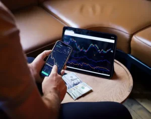 Person using a smartphone with a stock trading app while looking at a tablet displaying market charts on a coffee table, with cash nearby