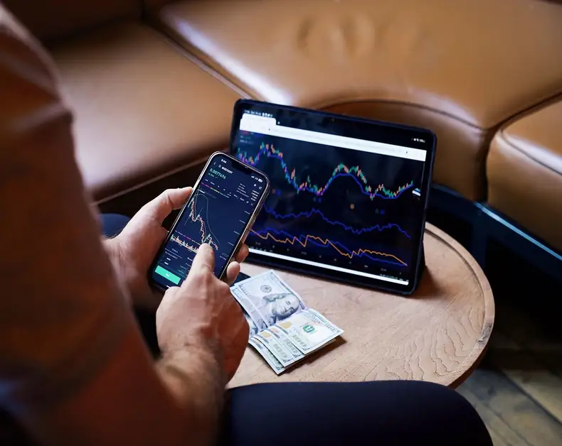 Person using a smartphone with a stock trading app while looking at a tablet displaying market charts on a coffee table, with cash nearby