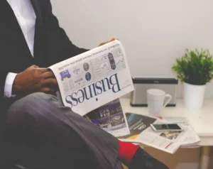 Person in a suit sits cross‑legged reading a business newspaper at a desk with a mug, plant, and phone nearby.