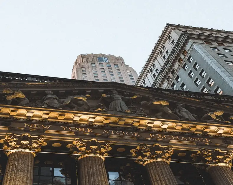View of the New York Stock Exchange facade with ornate sculptures and tall columns from street level, city buildings in the background.