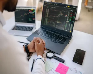 Trader analyzing stock charts on two laptops, hands clasped over a desk with a magnifying glass, a smartphone, and Buy/Hold/Sell notes nearby.